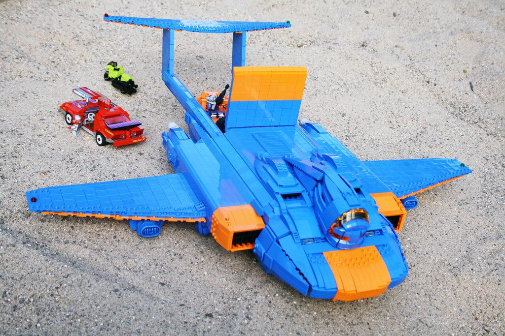 LEGO M.A.S.K. aircraft MOC with blue and orange jet, minifigure, and toy vehicles on concrete surface.