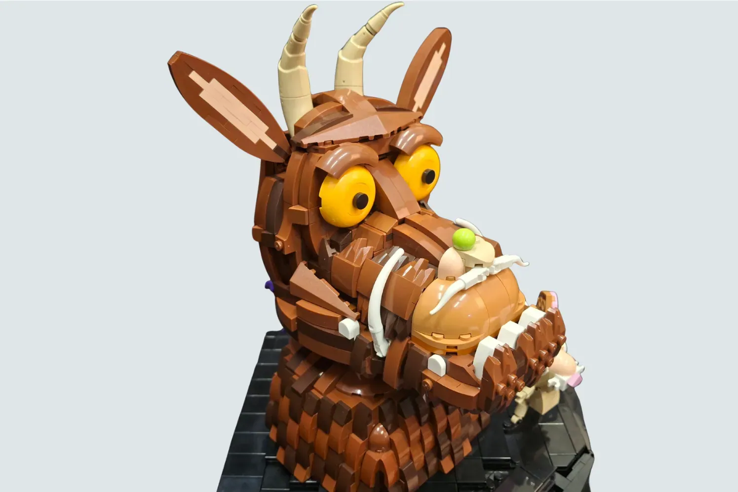 LEGO Gruffalo model with yellow eyes, tan horns, brown body, and small mouse figure on black base.