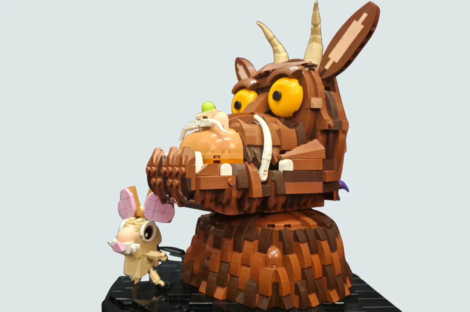 LEGO Gruffalo figure with large yellow eyes and horns, holding small mouse character on wooden base.