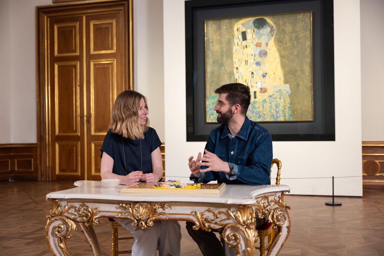Two people discussing LEGO Art Gustav Klimt set at ornate table with painting in background.