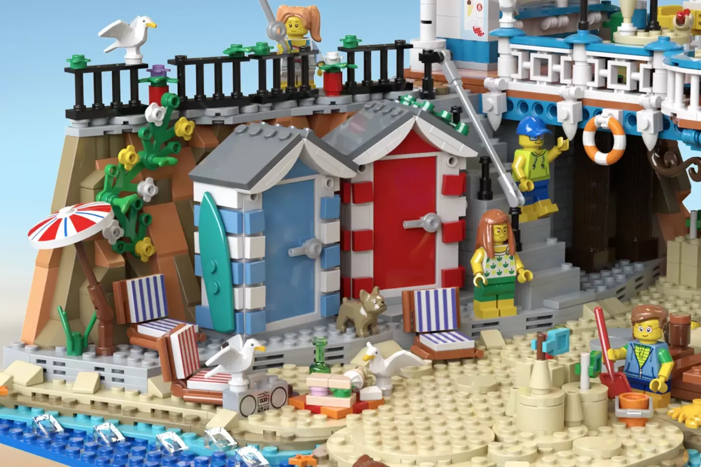 LEGO Victorian seaside pier with beach huts, minifigures, seagulls, beach chairs, and colorful buildings along sandy shore.