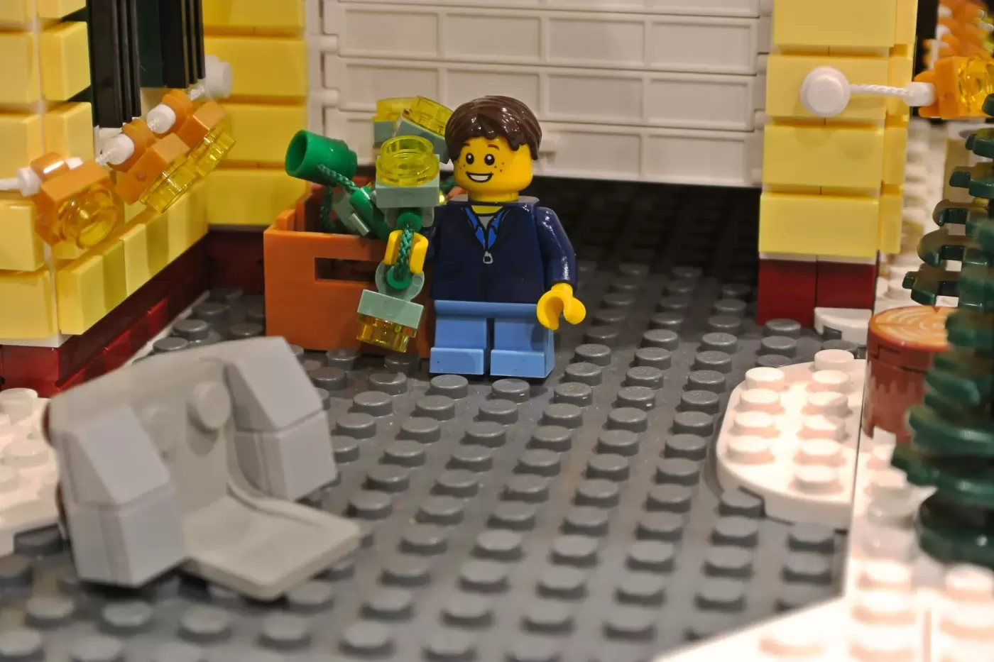 LEGO minifigure in blue jacket holding wrench inside decorated yellow house with garage and holiday lights.