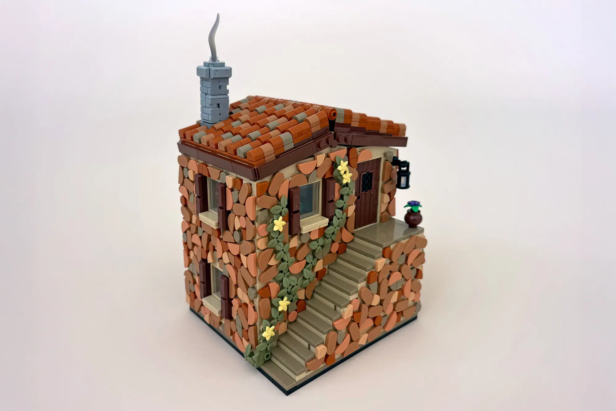 LEGO brick cottage with red tile roof, stone walls, climbing ivy, yellow flowers, chimney, and minifigure on steps.
