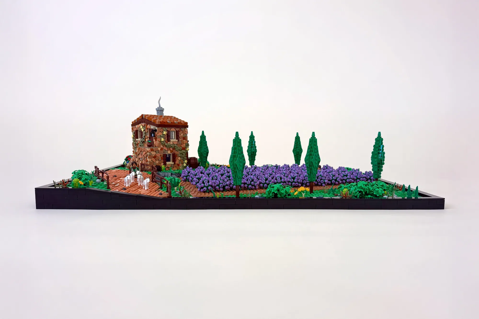 LEGO lavender field diorama with rustic stone tower, purple flower clusters, cypress trees, and minifigures on wooden base.