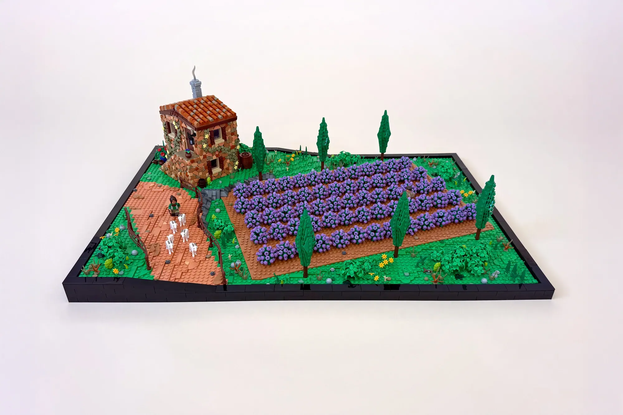LEGO lavender field diorama with stone cottage, purple flower rows, cypress trees, and minifigures on black base.