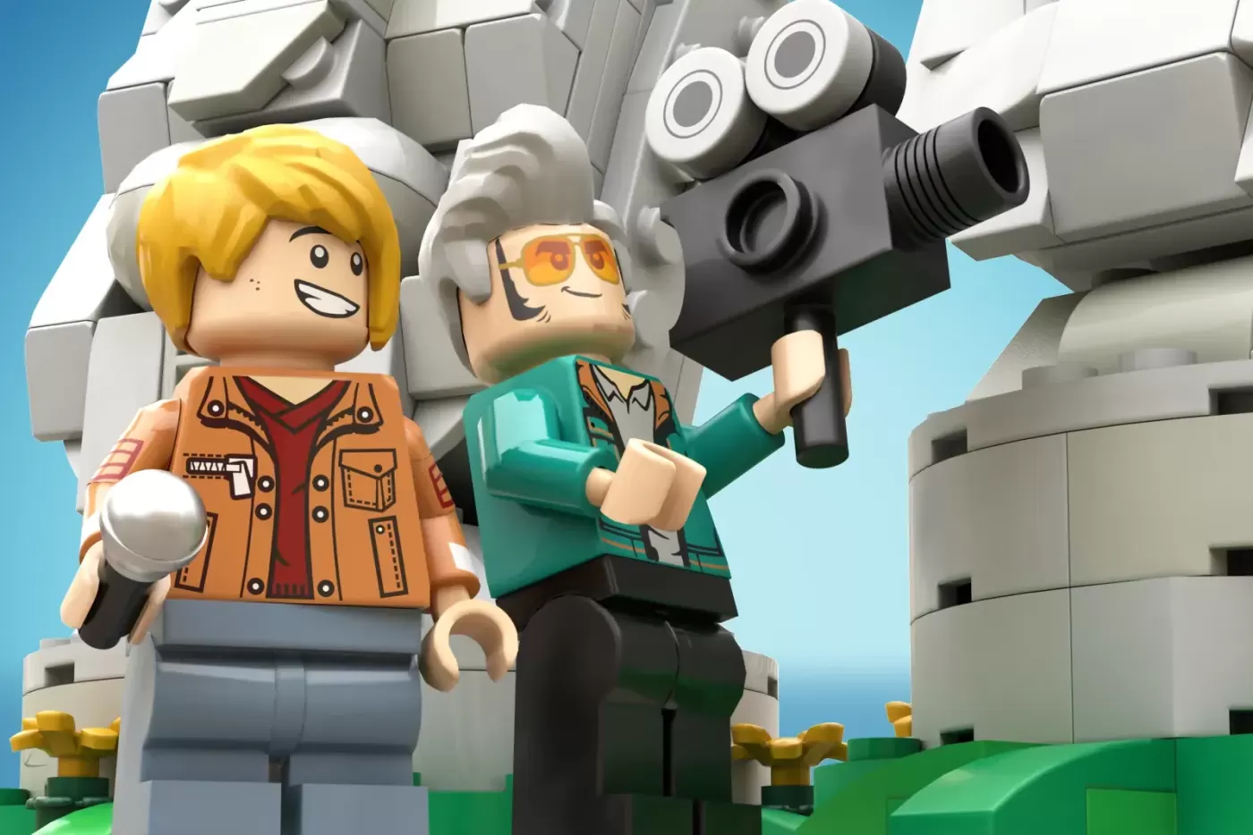 Two LEGO minifigures pose beside a large gray robot structure with camera equipment against a blue sky backdrop.