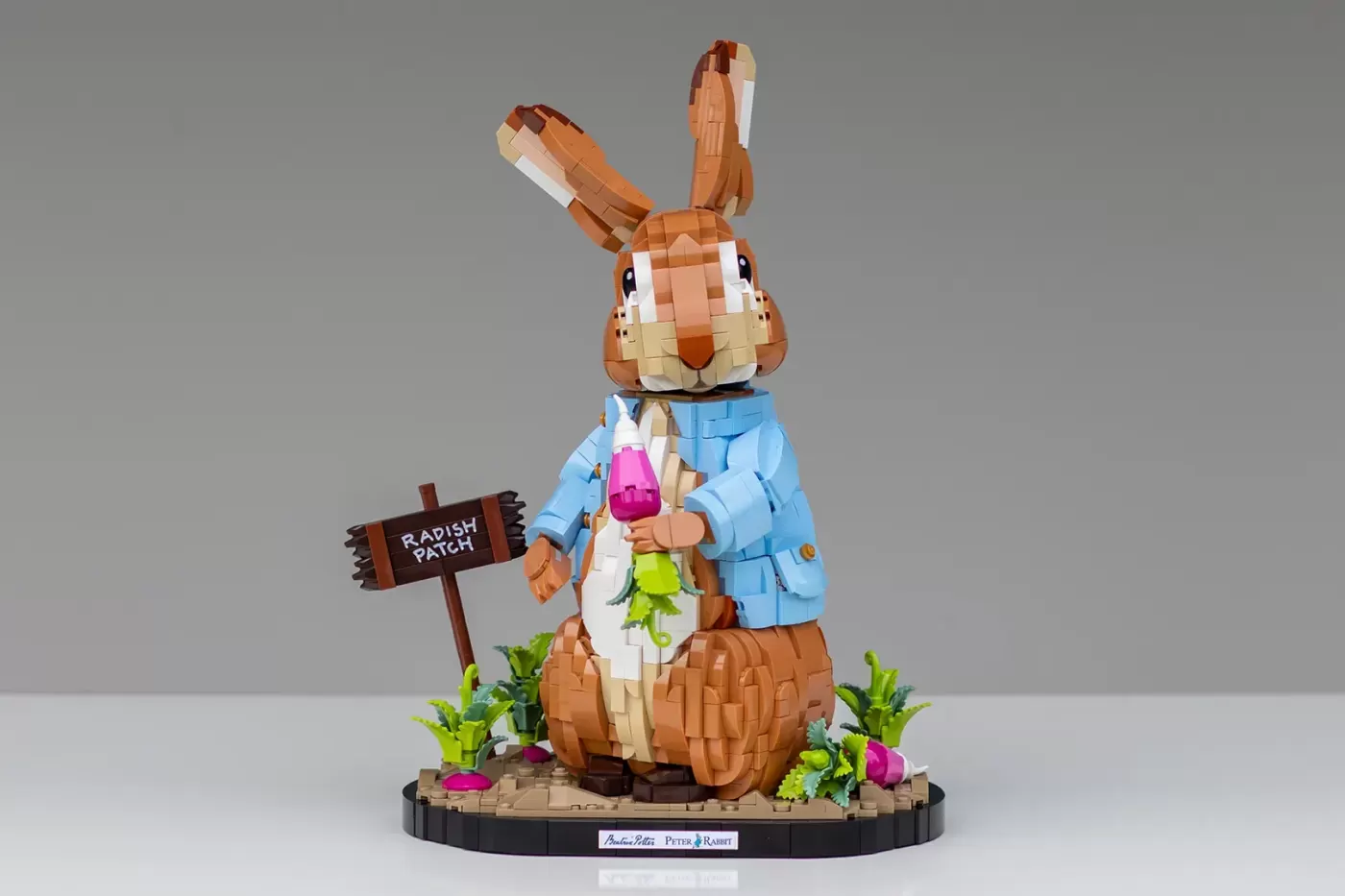 LEGO Peter Rabbit figure in blue jacket holding radish, standing on base with "Radish Patch" sign and vegetables.
