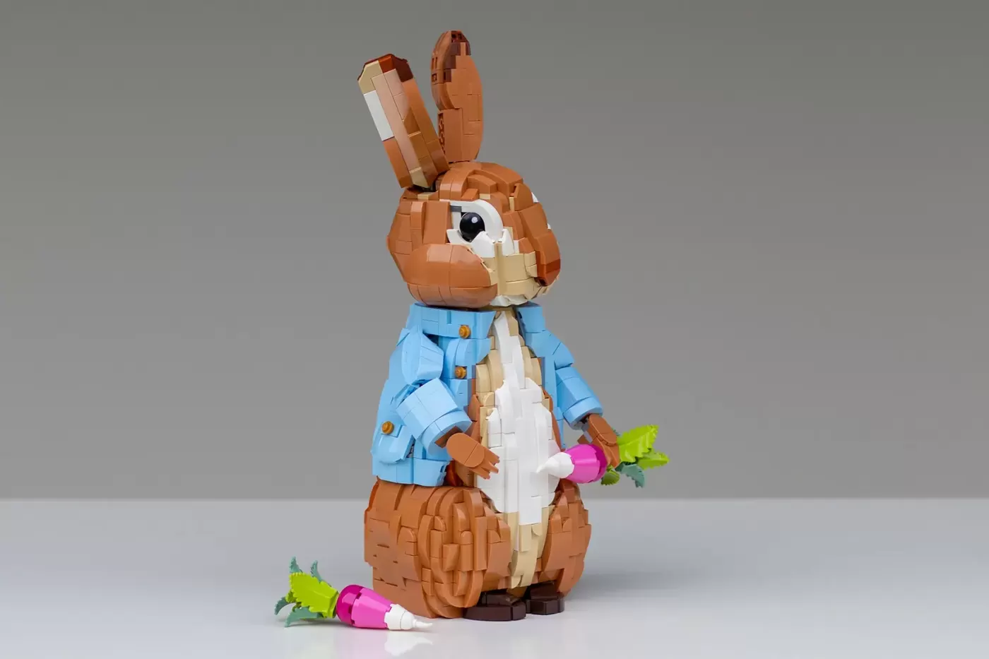 LEGO brick figure of Peter Rabbit in blue jacket holding a carrot, with pink flower accessory nearby.