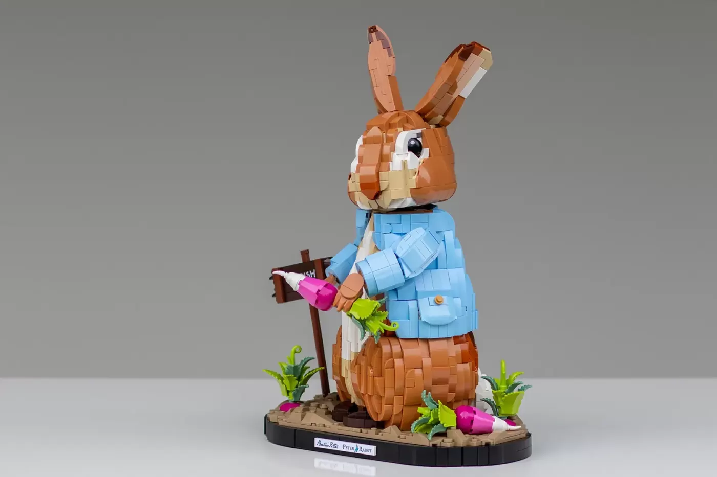LEGO Peter Rabbit figure in blue jacket holding vegetables, standing on base with garden plants and wooden sign.